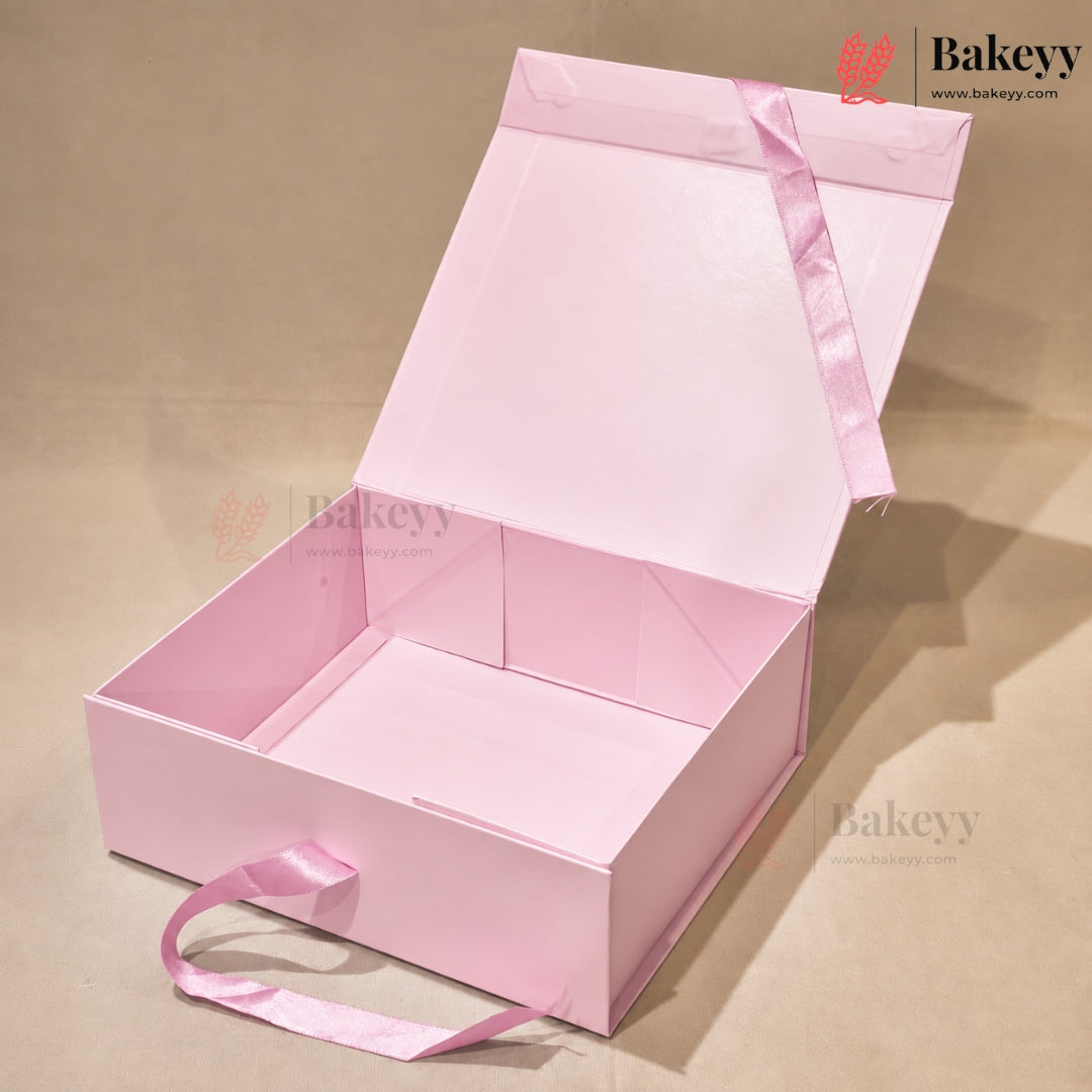 An open light pink cardboard gift box with a matching pink satin ribbon handle, photographed on a beige studio background. The box is shown from above at an angle, with the lid open and interior compartments visible.