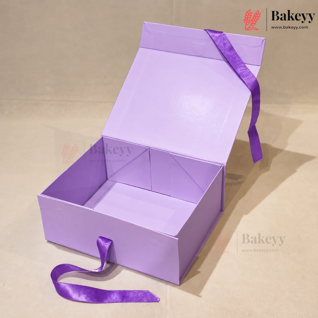 A lavender purple cardboard gift box with its lid open, featuring a matching purple satin ribbon tied around the handle and draped over the open top. The box is photographed on a neutral beige background with a visible Bakeyy brand watermark.