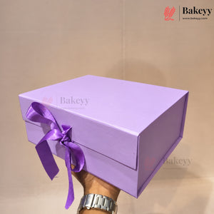 A lavender purple rectangular gift box with a matching purple ribbon bow, held by a hand with a silver watch against a plain beige background. The Bakeyy brand logo is visible in the top right corner.