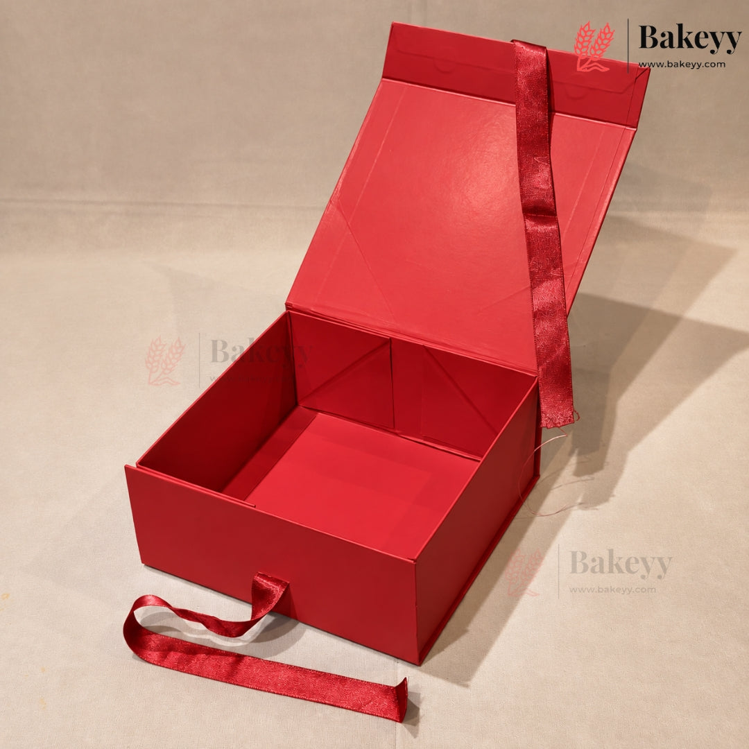 A red cardboard gift box with an open hinged lid, a red ribbon handle, and a matching red ribbon tied around the exterior, photographed on a light beige studio background.