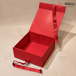 A red cardboard gift box with an open hinged lid, a red ribbon handle, and a matching red ribbon tied around the exterior, photographed on a light beige studio background.
