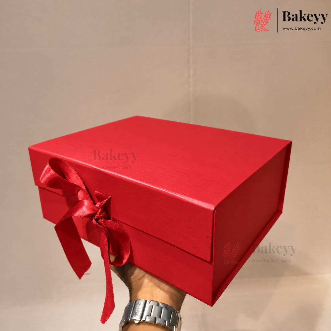 A bright red rectangular gift box with a matching red ribbon bow, held by a hand wearing a silver watch against a plain beige background. The Bakeyy logo is visible in the top right corner.