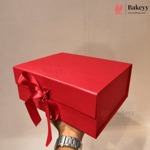 A bright red rectangular gift box with a matching red ribbon bow, held by a hand wearing a silver watch against a plain beige background. The Bakeyy logo is visible in the top right corner.