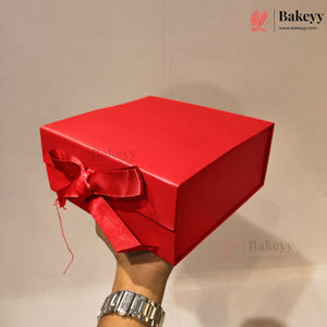 A hand holding a square red bakery gift box with a matching red satin ribbon bow, set against a plain beige background. The Bakeyy logo is visible in the top right corner.