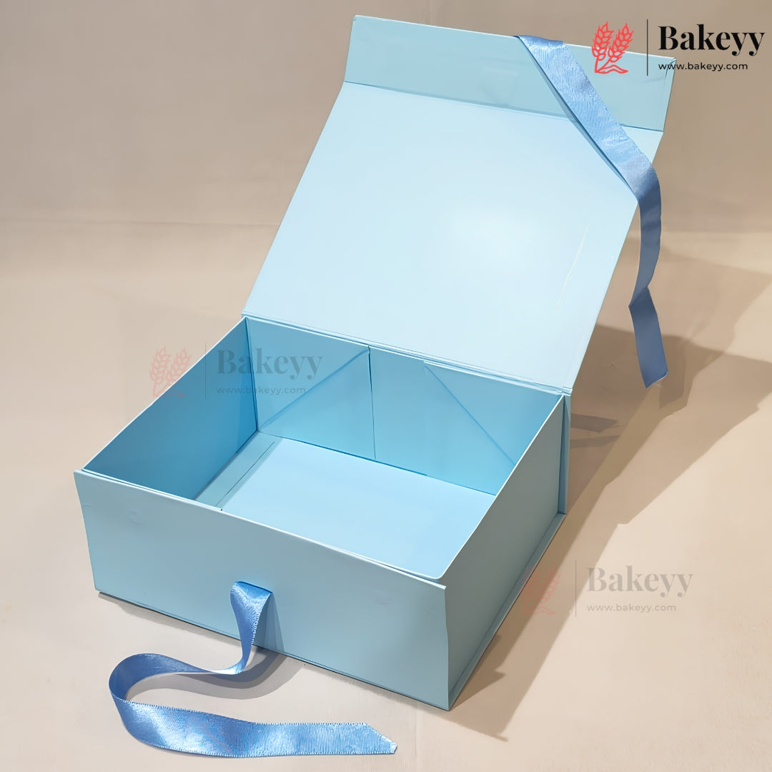 A light blue cardboard gift box with an open hinged lid, featuring a matching light blue satin ribbon tied around the lid and a small loop on the front side. The box is photographed on a plain beige studio background with a Bakeyy watermark and logo visible.