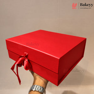 A bright red rectangular gift box with a matching red ribbon bow, held by a hand wearing a silver watch against a plain beige background. The Bakeyy logo is visible in the top right corner.