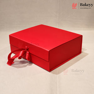 A red rectangular bakery gift box with a matching red satin ribbon tied in a bow, photographed on a light beige background. The Bakeyy brand logo is visible in the top right corner.