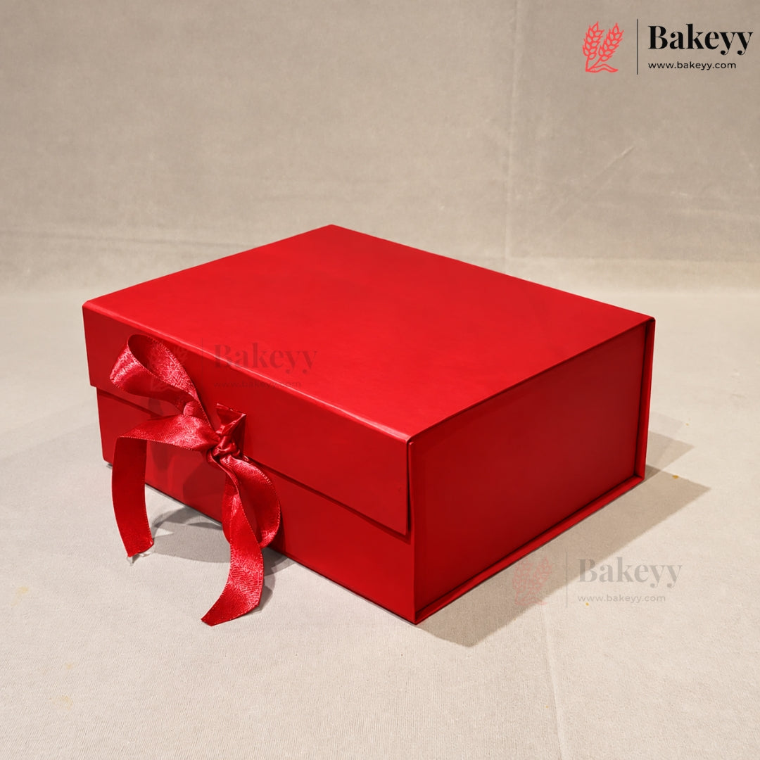 A red rectangular gift box with a matching red ribbon bow on the front, photographed on a light beige textured background. The Bakeyy logo is visible in the top right corner.