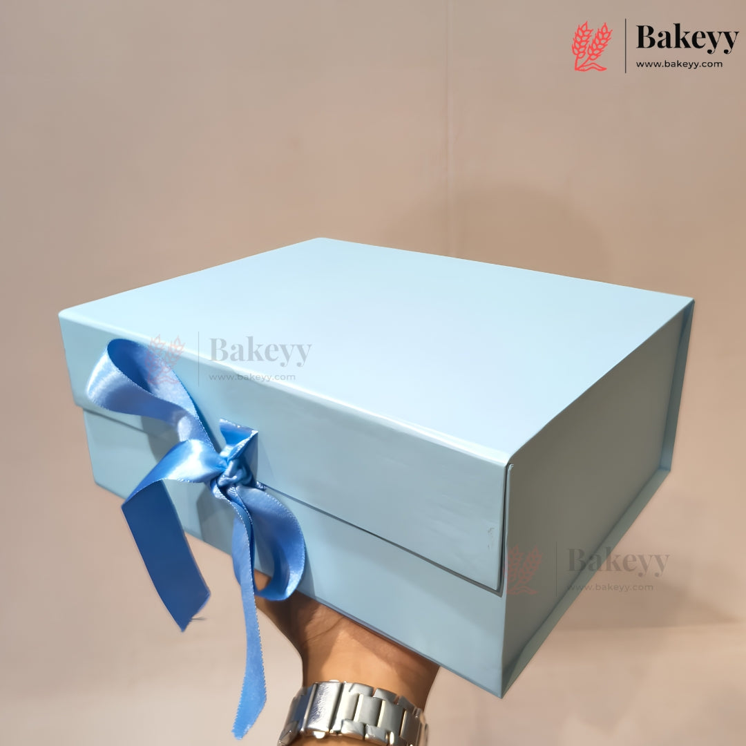 A light blue rectangular gift box with a matching blue satin ribbon bow on the lid, held by a hand wearing a silver watch against a plain beige background. The Bakeyy logo is visible in the top right corner.