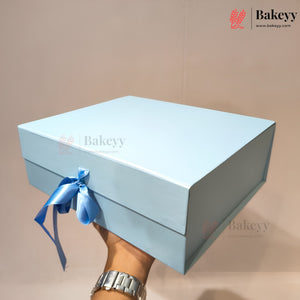 A light blue rectangular gift box with a matching blue ribbon bow, held by a hand wearing a silver watch against a plain beige background. The Bakeyy logo is visible in the top right corner.