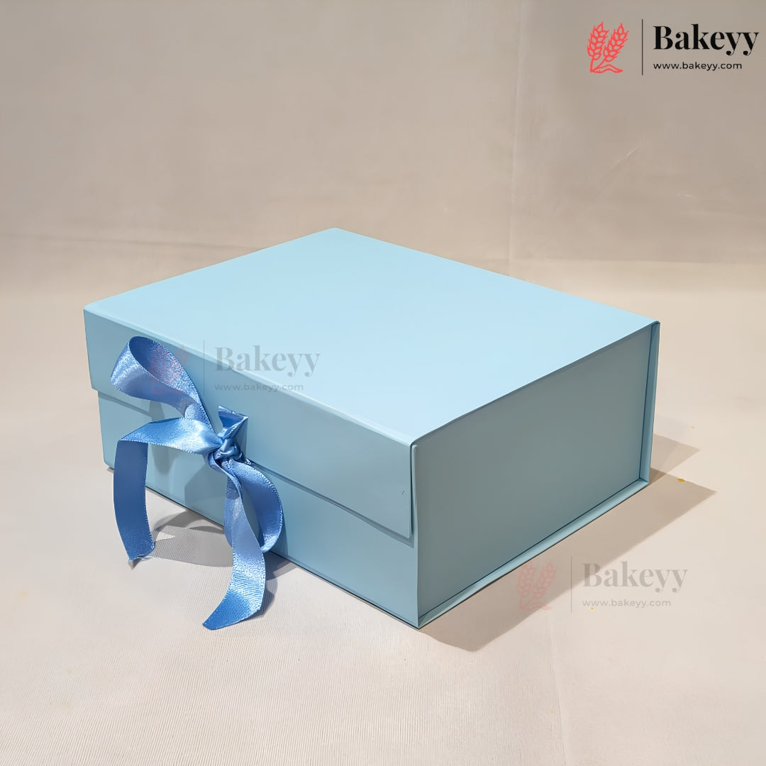 A light blue rectangular gift box with a matching blue satin ribbon tied in a bow on the front, photographed on a plain beige studio background. The Bakeyy logo appears in the top right corner.