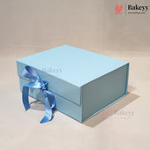A light blue rectangular gift box with a matching blue satin ribbon tied in a bow on the front, photographed on a plain beige studio background. The Bakeyy logo appears in the top right corner.