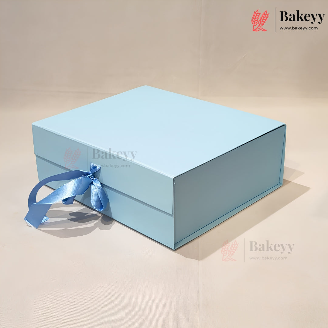 A light blue rectangular gift box with a matching blue ribbon tied in a bow on the front, photographed on a plain beige studio background. The Bakeyy logo appears in the top right corner.