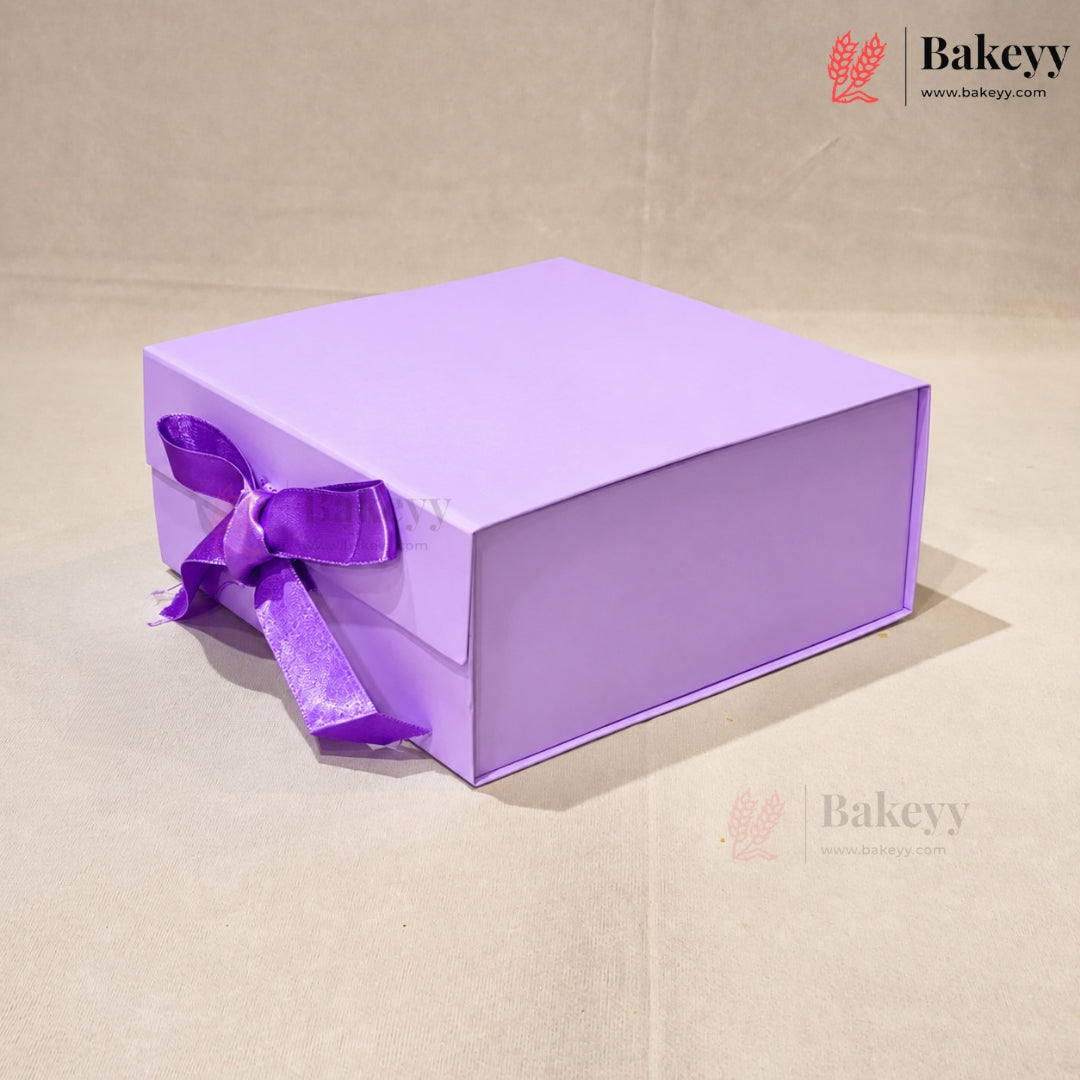 A lavender purple square gift box with a matching purple satin ribbon tied in a bow on the front, set against a neutral beige background. The Bakeyy brand logo is visible in the top right corner.