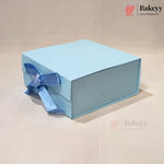 A light blue square gift box with a matching blue ribbon bow, photographed on a plain beige background. The Bakeyy logo is visible in the top right corner.