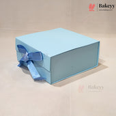 A light blue square gift box with a matching blue ribbon bow, photographed on a plain beige background. The Bakeyy logo is visible in the top right corner.