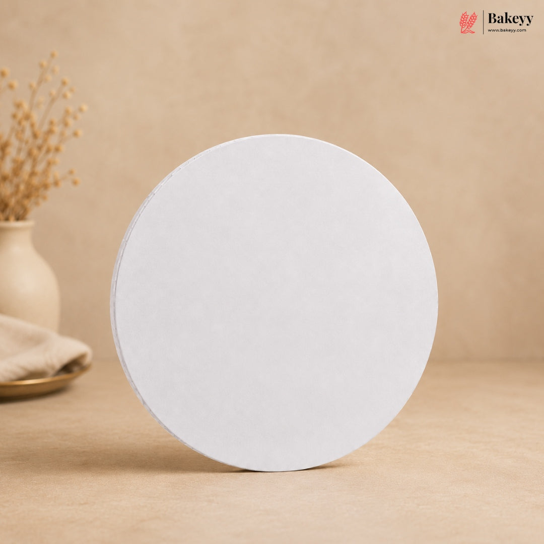 A round white cake stand or display riser standing upright on a beige surface against a warm beige background, with a small vase of dried stems in the left background and a Bakeyy logo in the top right corner.