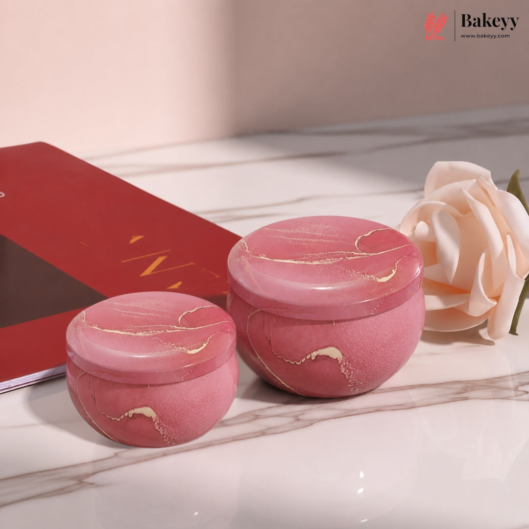 Two pink marbled cosmetic jars with gold veining, displayed on a white marble surface next to a red book and a pale pink rose. A Bakeyy logo is visible in the top right corner.