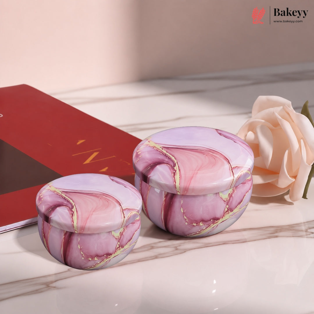Two round pink marble-patterned lidded containers with gold veining, displayed on a marble surface next to a red book and a pale peach rose. A Bakeey logo watermark is visible in the top right corner.