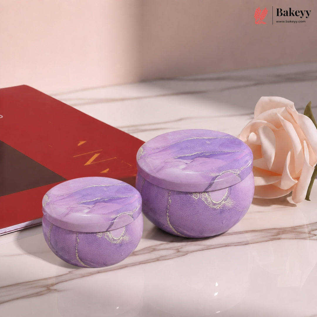Two round purple marbled cosmetic jars with lids, displayed on a marble surface next to a red book and a pale pink rose.