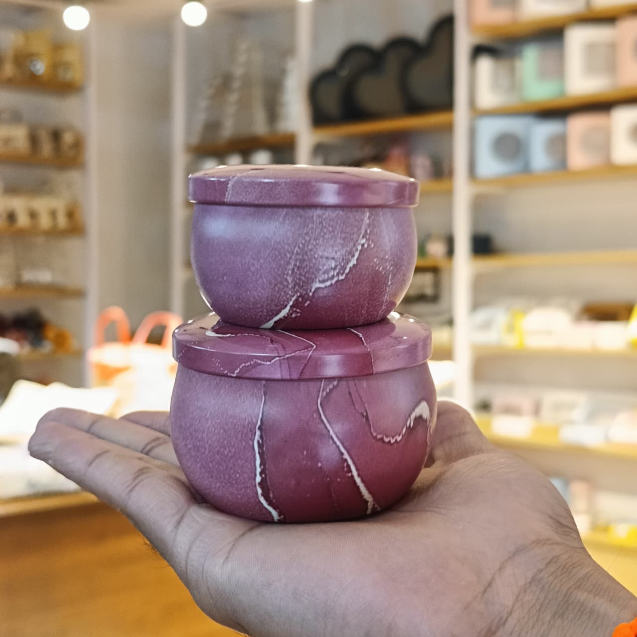 A hand holding two stacked round purple stone containers with lids, featuring natural white veining, set against a blurred background of a retail shop.