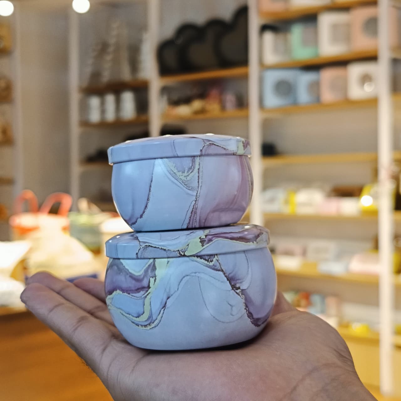 Two small round lidded jars stacked on a person's hand, featuring a lavender and purple marble pattern with gold veining, set against a blurred background of a shop interior.