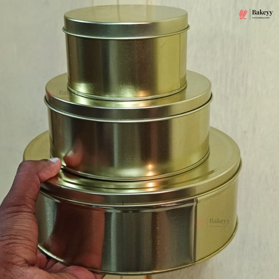 Three nested round metal tins with lids in a brushed gold finish, held by a hand against a light beige wall. The Bakeyy logo is visible in the top right corner.