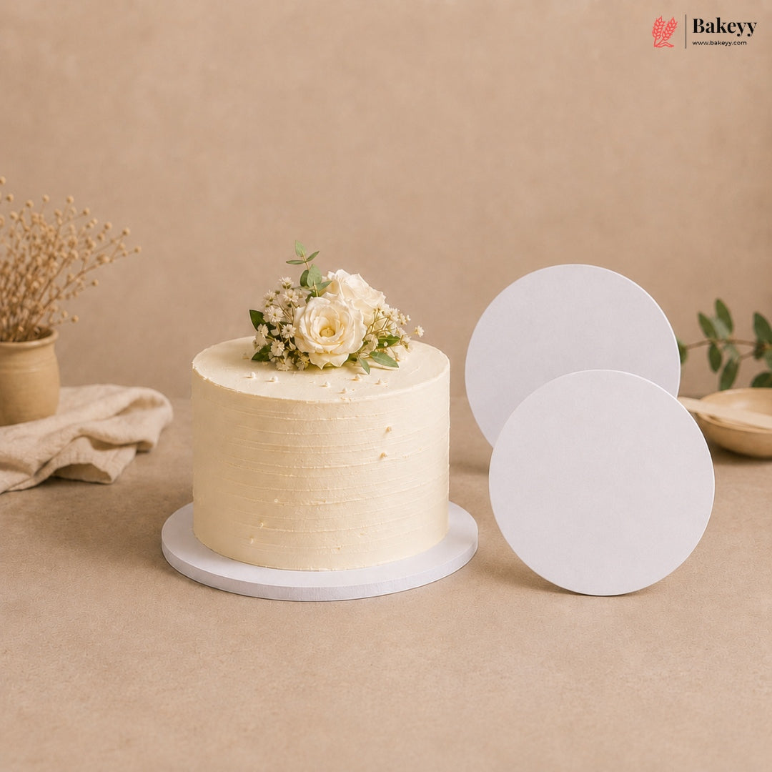 A cream-colored frosted cake with white flowers and green leaves sits on a round white cake stand, with two blank white round cake stands displayed beside it on a beige background. The Bakeyy logo is visible in the top right corner.