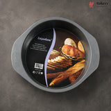 A round non-stick baking pan with side handles, shown from above on a dark gray textured surface. The inside of the pan has a large black product label featuring bread imagery and the brand name 'Cupcake Pan'.