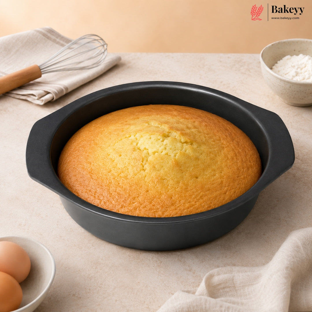 A round black cake pan with side handles containing a golden-brown baked cornbread or cake, styled on a beige countertop with baking ingredients and utensils nearby. The Bakey brand logo appears in the top right corner.