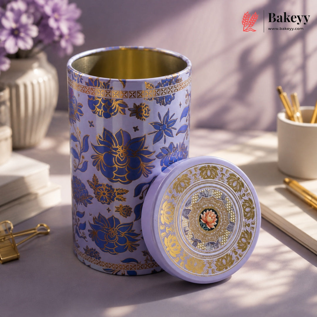 A decorative cylindrical metal tea tin with a matching lid, featuring a lavender and gold floral pattern with Chinese characters, displayed on a styled desk surface.