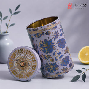 A decorative cylindrical metal tea or coffee canister with its lid placed beside it. The canister has a lavender-purple exterior with blue floral motifs and gold detailing, and a gold interior.