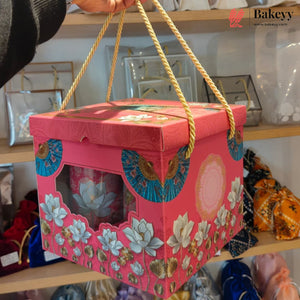 A pink decorative gift box with floral and fan patterns, held by a hand with gold rope handles, set against a blurred store background.