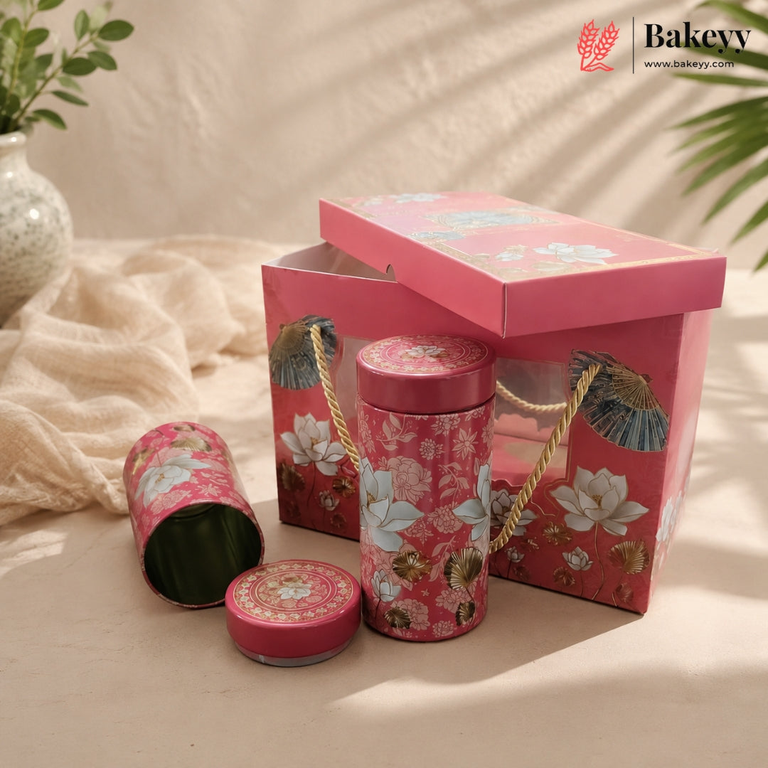 A pink floral bakery gift set featuring a rectangular box with a hinged lid, a cylindrical tin with rope handles, a small round tin, and an open tin showing green contents, styled on a beige fabric surface with soft shadows and a Bakeyy logo in the top right corner.