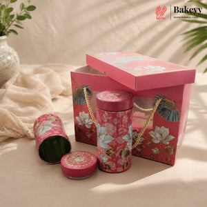 A pink floral bakery gift set featuring a rectangular box with a hinged lid, a cylindrical tin with rope handles, a small round tin, and an open tin showing green contents, styled on a beige fabric surface with soft shadows and a Bakeyy logo in the top right corner.