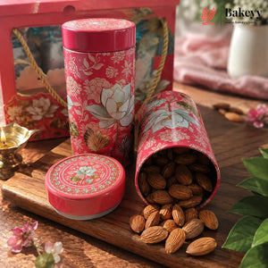 A pink floral metal tin container for baking or tea, shown closed and open with almonds spilling out on a wooden surface. A matching gift box is visible in the background, along with decorative flowers, a small brass lamp, and green leaves.