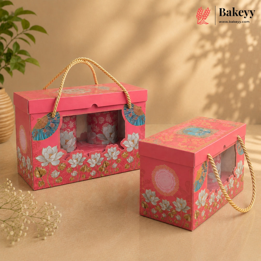 Two pink decorative bakery gift boxes with floral prints and rope handles, shown on a warm beige background with a plant and dried flowers as props. The Bakeyy logo appears in the top right corner.