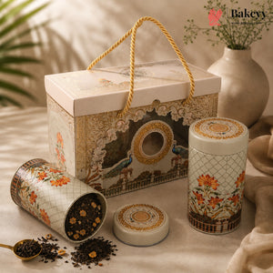 A decorative tea gift set with a rectangular illustrated box and two matching cylindrical tins, shown on a draped fabric surface with loose tea leaves and a spoon. The packaging features ornate floral and peacock artwork in beige, gold, orange, and brown tones.