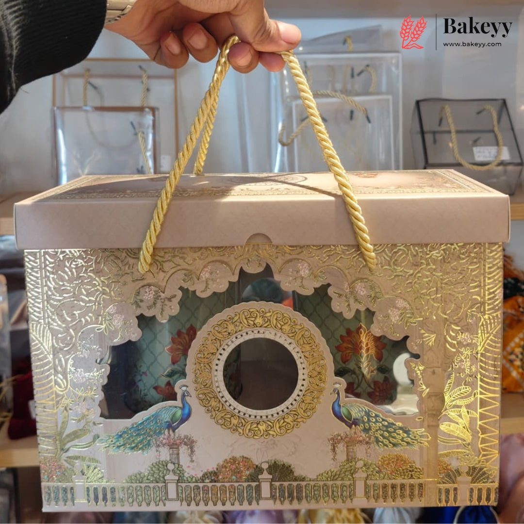 An ornate rectangular gift box with a lid, decorated with gold foil patterns, peacock illustrations, and a central oval window. The box has twisted rope handles and is being held by a hand in a retail setting.