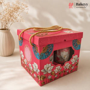 A pink decorative gift box with a rope handle, floral and fan pattern, and a clear window showing a pink lotus cup inside. A beige vase with dried stems sits in the background on a warm cream surface.