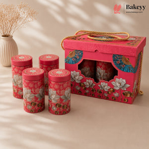 A pink tea gift set with four cylindrical floral tins and a matching decorative box, styled on a beige surface with a vase of dried flowers in the background. The Bakeyy logo is visible in the top right corner.
