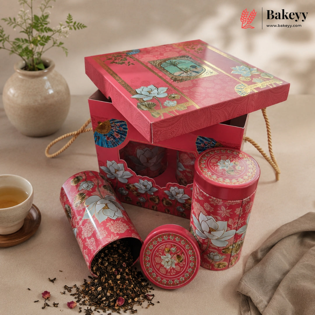 A red floral tea gift set with a matching box, two round metal tins, and loose tea scattered on a beige surface. A small cup of tea, a vase with greenery, and a folded fabric cloth are arranged around it.