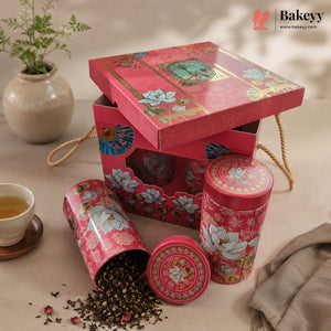 A red floral tea gift set with a matching box, two round metal tins, and loose tea scattered on a beige surface. A small cup of tea, a vase with greenery, and a folded fabric cloth are arranged around it.