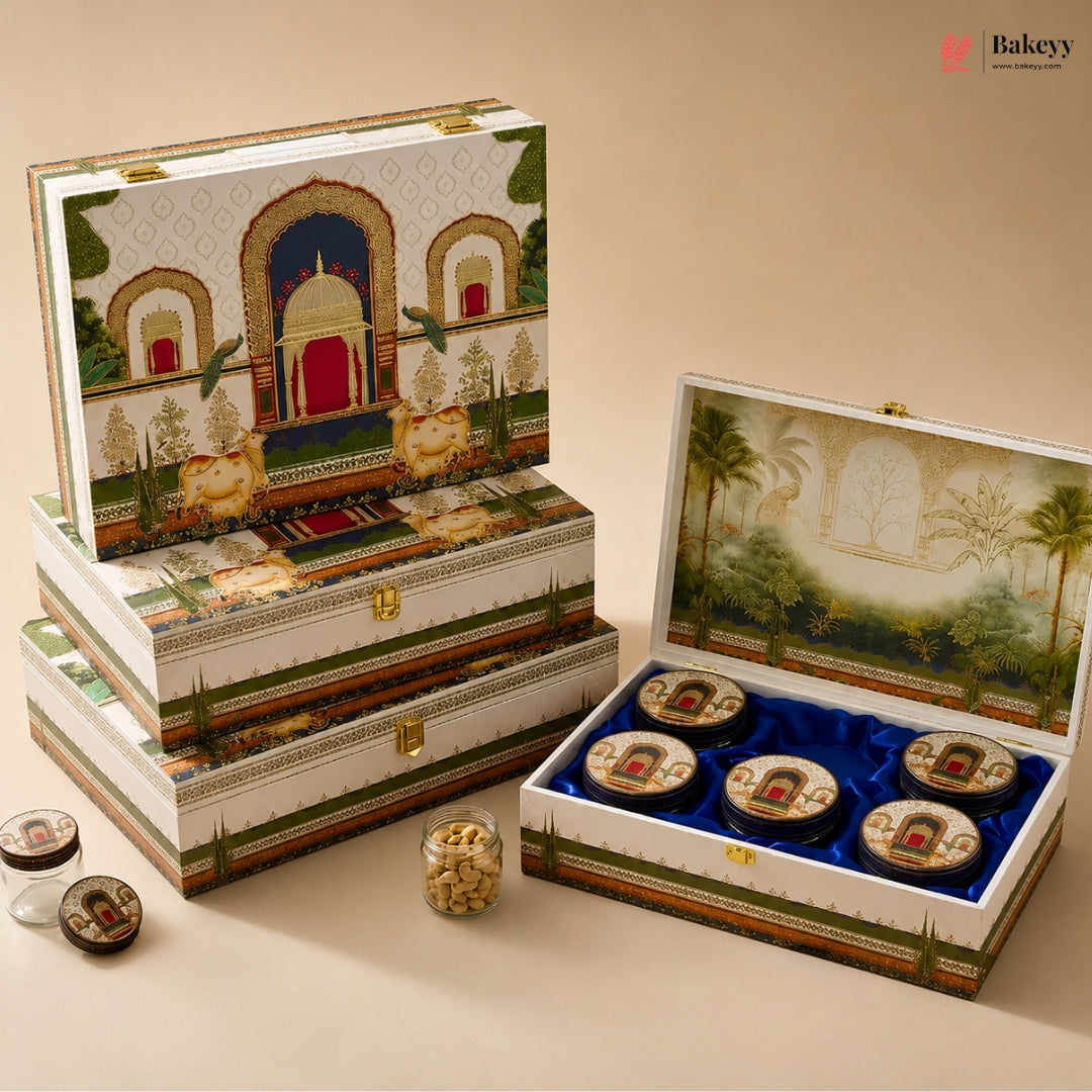An ornate gift box set with traditional Indian-style artwork, shown closed and open to reveal round packaged items on a blue velvet insert. The boxes feature detailed illustrations of domed architecture and scenic landscapes, with small jars of snacks placed beside them on a warm beige background.