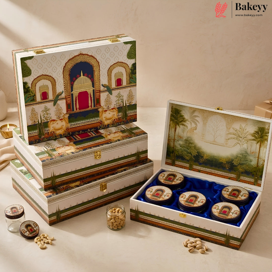 An ornate bakery gift set displayed on a beige surface, featuring stacked rectangular boxes with detailed architectural illustrations and one open box containing four round pastries on a blue velvet insert. Small jars of nuts and candies are placed nearby.