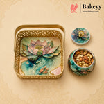 A decorative gold metal gift tray with a lotus pond design, shown alongside a matching lidded container and a small jar of nuts. The Bakeyy brand logo is visible in the top right corner.