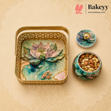 A decorative gold metal gift tray with a lotus pond design, shown alongside a matching lidded container and a small jar of nuts. The Bakeyy brand logo is visible in the top right corner.