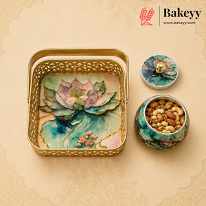 A decorative gold metal gift tray with a lotus pond design, shown alongside a matching lidded container and a small jar of nuts. The Bakeyy brand logo is visible in the top right corner.