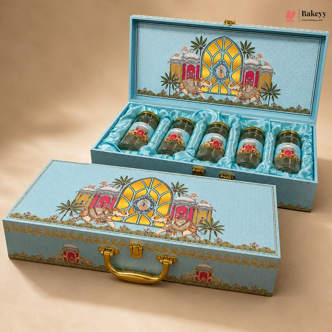 A decorative turquoise gift box set with five small jars arranged inside an open box and one matching closed box in front. The boxes feature a colorful architectural illustration with pink carriages, palm trees, and gold details, and the jars have gold lids.