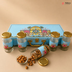 A gourmet gift set of five glass jars with gold lids and blue decorative labels, arranged in front of a matching blue illustrated storage box. One jar is tipped over with almonds spilling out onto a beige surface, with a small glass dish of almonds in the foreground.